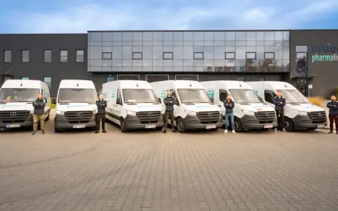 Sześciu mężczyzn w uniformach stoi przed rzędem białych vanów z logo "pharmalink". To zespół odpowiedzialny za transport leków, profesjonalny przewoźnik.