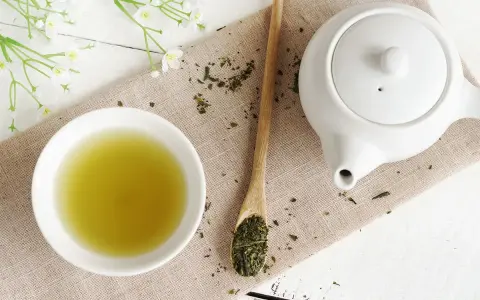Relaksująca zielona herbata na noc. Filiżanka naparu, biały czajniczek i drewniana łyżka z liśćmi herbaty.