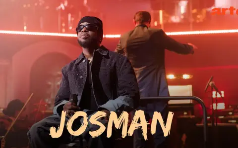 Josman, francuski rap artysta, siedzi na scenie z mikrofonem, w tle orkiestra i dyrygent.