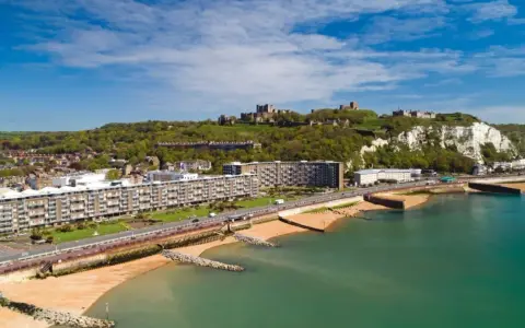 Vista aerea del porto di Dover con il castello in cima alla scogliera bianca. Edifici moderni e spiagge sabbiose si affacciano sul mare turchese.