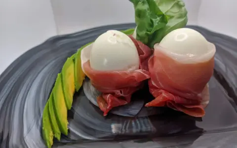 Due uova di quaglia sode avvolte in prosciutto crudo, servite con fette di avocado e foglie verdi su un piatto scuro.