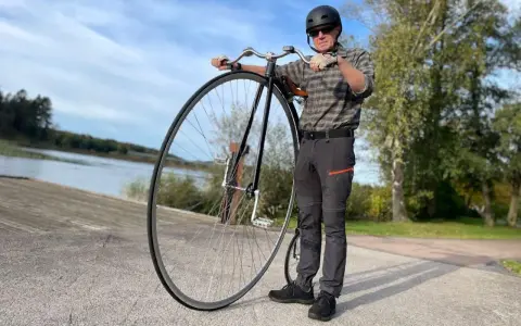 Man med hjälm och solglasögon står bredvid en cykel med stort framhjul vid en sjö.