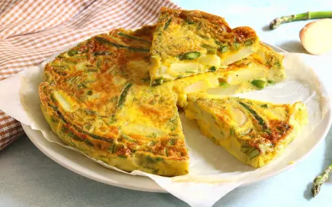 Una gustosa frittata di asparagi, con patate e uova dorate, tagliata a fette su un piatto bianco.
