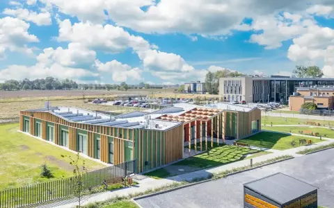 Nowoczesny budynek użyteczności publicznej z zielonymi fasadami i drewnianymi elementami, otoczony trawnikami i parkingiem.