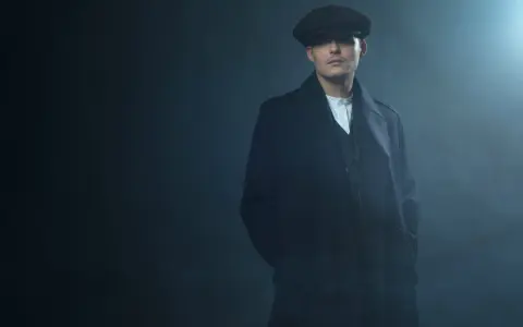 Młody mężczyzna w czapce i płaszczu, z charakterystyczną fryzurą w stylu Peaky Blinders, stoi w zadymionym mroku.