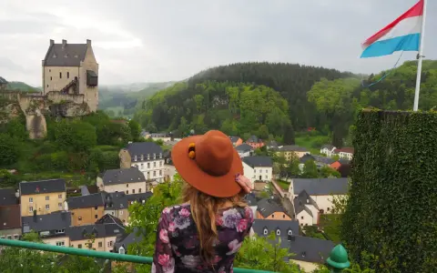 Mujer con sombrero marrón admira un castillo y un pueblo, con la bandera de Luxemburgo ondeando. ¡Un paisaje digno de un buen queso Muenster!