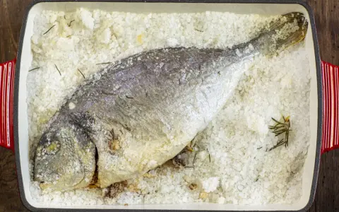 Orata al sale, intera e dorata, adagiata su un letto di sale grosso e rosmarino in una teglia rossa.