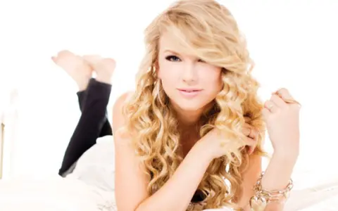 Taylor Swift w stylu "Speak Now" z burzą blond loków, w czarnych legginsach, leży na łóżku.