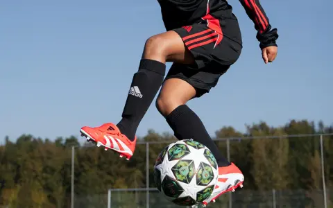 Barn i luften med fotboll. Svarta strumpor med Adidas-logga och benskydd fotboll barn storlek. Orange fotbollsskor med vita detaljer.