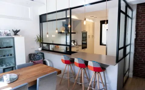 Tabique cocina semiabierta al salón con barra y taburetes coloridos. La cocina moderna se integra al salón a través de un panel de cristal y metal.