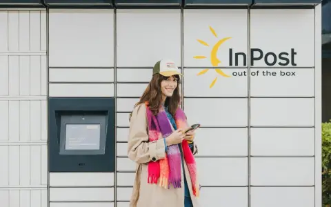 Uśmiechnięta dziewczyna z telefonem w ręku przy paczkomacie InPost. Szybki kurs obsługi, by odebrać przesyłkę.