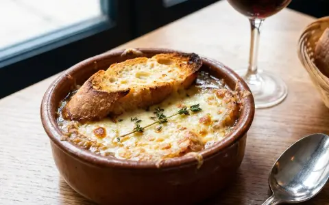Un cuenco de sopa de cebolla gratinada con pan tostado y queso derretido, adornado con tomillo. Una copa de vino tinto y una cuchara completan la escena.