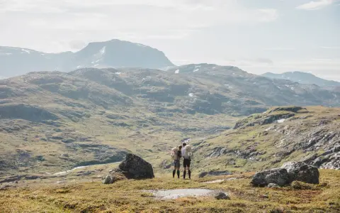 Två vandrare med bamm utrustning står på en bergssluttning med vidsträckt utsikt över fjällandskap.
