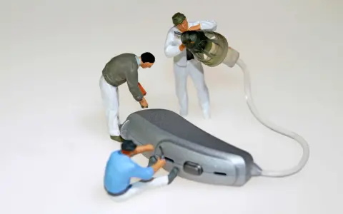 Miniaturowi pracownicy zajmują się naprawą aparatów słuchowych. Jeden z nich przykręca element, drugi czyści słuchawkę, a trzeci pracuje nad aparatem.