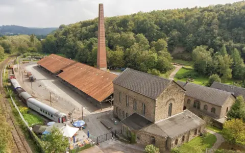 Alte Zechengebäude der Zeche Nachtigall mit einem langen roten Dach, einem hohen Backsteinschornstein und einem historischen Zug.
