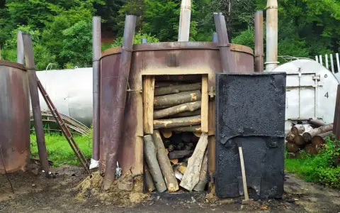 Wypalanie węgla drzewnego w piecu. Drewno ułożone w otwartym palenisku, obok czarne drzwi i inne metalowe konstrukcje.