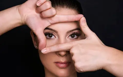 Mani incorniciano il volto di una donna, creando un effetto di profondità. Si può vedere un alone nell'occhio, quasi un riflesso di luce.