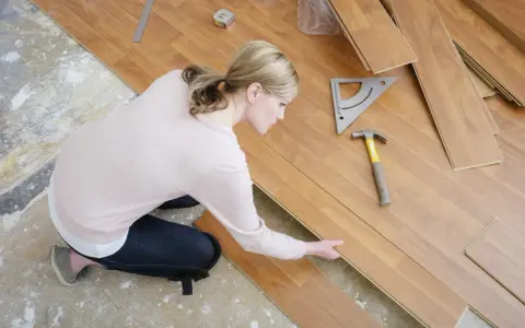 Frau kniet und verlegt Laminat. Werkzeuge wie Hammer und Winkel liegen bereit.