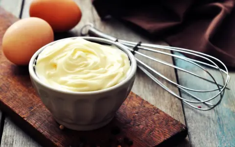 Un bol de mayonnaise onctueuse, des œufs et un fouet, prêts à faire une mayonnaise maison.