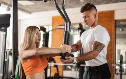 Kobieta ćwiczy na siłowni pod okiem trenera. On pokazuje jej prawidłowe wykonanie ćwiczenia, co jest kluczowe, gdy stawiasz pierwsze kroki i poznajesz **trener personalny wymagania**.