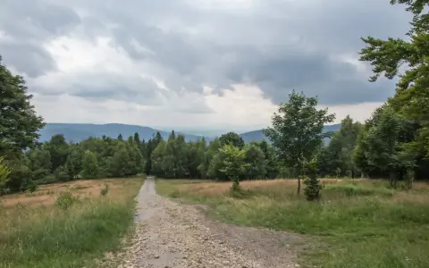Kamienista ścieżka prowadzi przez trawiaste zbocze ku lasowi. W oddali widać pasma górskie pod pochmurnym niebem. To miejsce, gdzie można poczuć się jak na hrobaczej łące.