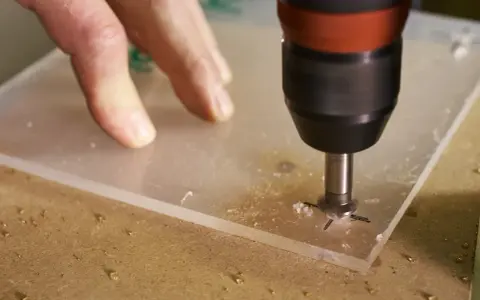 A drill bit is used to drill through acrylic. A hand guides the acrylic sheet as the drill creates a hole, showing it's possible to drill through acrylic.