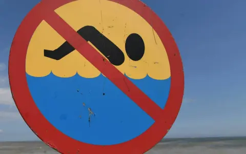Znak zakazu kąpieli na plaży. Zamknięte kąpieliska nad morzem, mimo pięknej pogody i widocznej w oddali osoby w wodzie.