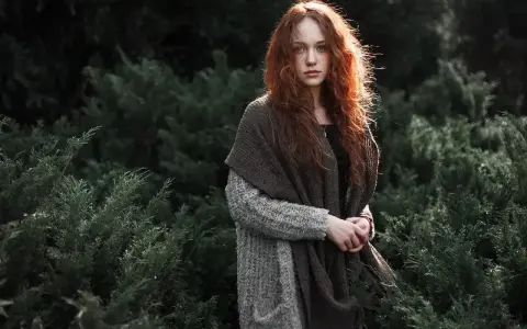 Rudowłosa dziewczyna w grubym swetrze i szalu, otoczona ciemną zielenią. Jej spojrzenie jest spokojne, a cała scena ma **low-key** klimat.