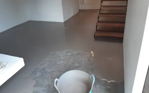 Pavimento continuo de hormigón gris recién aplicado, con una escalera de madera al fondo y un cubo con material.
