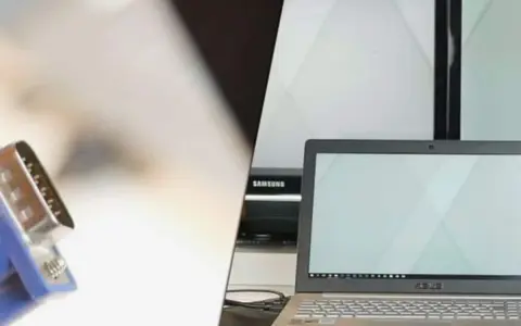 Ein VGA-Kabel wird verwendet, um einen Laptop mit einem Bildschirm zu verbinden. Mehrere Monitore sind im Hintergrund zu sehen.