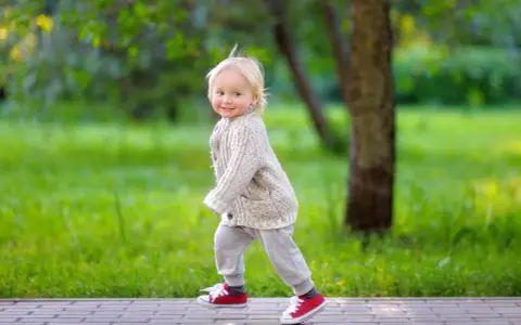 Ein fröhliches Kind, ca. 2,5 Jahre alt, mit blonden Haaren und einem Strickcardigan, läuft auf einem Weg im Park.