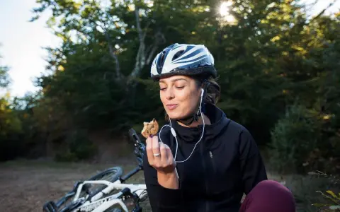 Kobieta w kasku i słuchawkach je ciastko, obok leży rower. To przykład zdrowego odżywiania i aktywnego stylu życia, czyli zbilansowana dieta.
