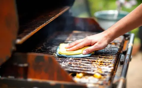 Main d'une personne nettoyant un barbecue très encrassé avec une éponge jaune et bleue.