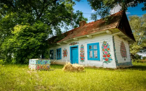 Malowany dom w Zalipiu, z niebieskimi oknami i drzwiami, ozdobiony kwiatami. Obok studnia i snop siana. Cennik wizyty w tym malowniczym miejscu jest dostępny dla zwiedzających.