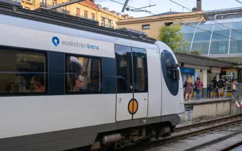 A modern Euskotren train at San Sebastian train station. Passengers wait on the platform under a glass-covered canopy.