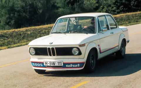 Białe BMW E20 z kolorowymi pasami i napisem "turbo" na błotniku, z kierowcą za kierownicą, jedzie po asfaltowej drodze.