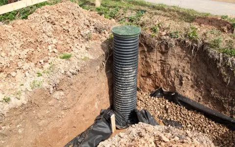 Schemat drenażu działki: pionowa rura drenarska w wykopie, otoczona kamieniami i geowłókniną.