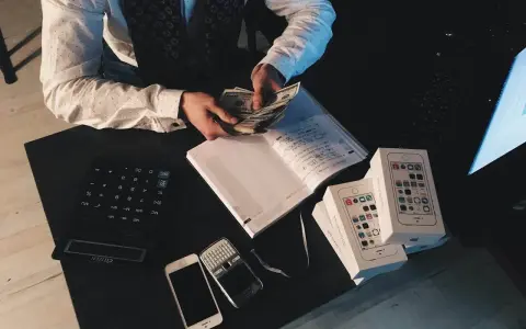Mężczyzna liczy pieniądze przy biurku z kalkulatorem, notatnikiem, telefonami i pudełkami po iPhone'ach. To może być jego sposób na zarządzanie finansami, być może w kontekście aml.