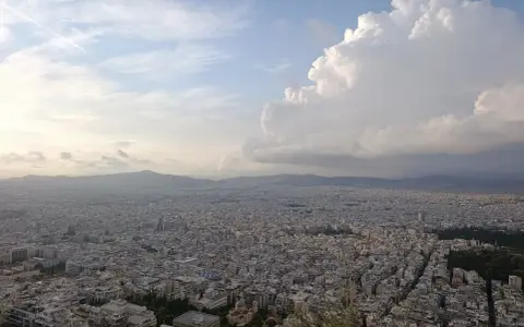 Rozległa panorama Aten z wzgórza Lycabettus. Miasto rozciąga się aż po horyzont, a nad nim piętrzą się chmury.