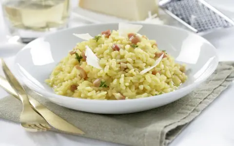 Un cremoso arroz a la milanesa, adornado con láminas de queso parmesano y hierbas frescas, servido en un plato blanco.