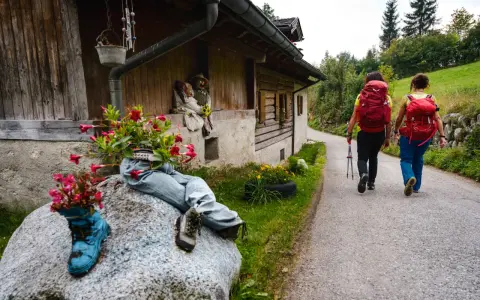 Due escursioniste affrontano il cammino San Vili, con zaini rossi e bastoncini. La strada, tra case rurali e verde, suggerisce una certa difficoltà.