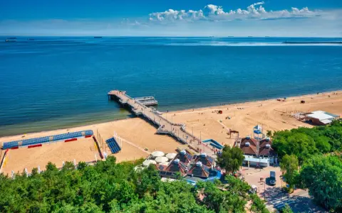 Idealne miejsce, gdzie na wakacje w sierpniu: piaszczysta plaża, molo, morze i zielone drzewa.