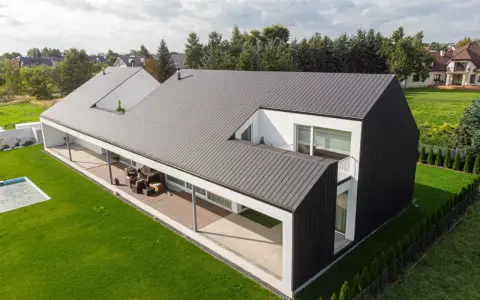 Nowoczesny dom z czarną elewacją i dachem krytym blachą rąbek. Otoczony zielonym ogrodem z basenem.
