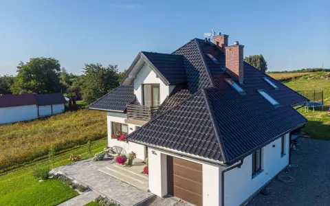 Nowoczesny dom z czarnym dachem, odpornym na warunki atmosferyczne, klasa korozyjności C4. Otoczony zielenią i zadbanym podjazdem.