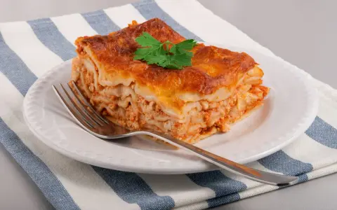 Une part de lasagne maison, dorée et appétissante, garnie d'une feuille de persil, sur une assiette blanche avec une fourchette.