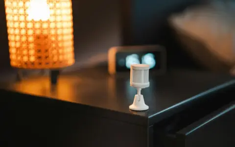 Sensor de movimiento blanco sobre mesita de noche, junto a lámpara y reloj. Ideal para saber como dejar fijo un sensor de movimiento.