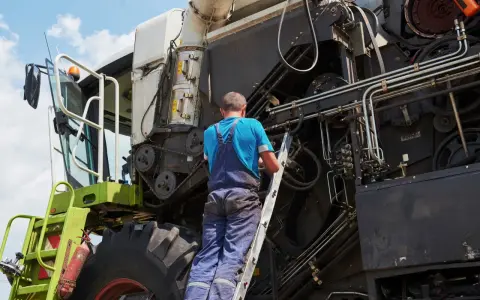 Mechanik na drabinie naprawia kombajn. Co to jest tuz? To potężna maszyna rolnicza, która zbiera plony.