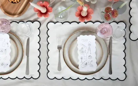 A table setting with a gold-colored tablecloth, white placemats with black scalloped edges, plates, cutlery, glasses, and a menu with illustrations.