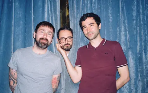 Trois hommes posent devant un rideau bleu. L'un d'eux, au centre, est un groupe musique espagnol année 2000.