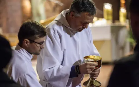 Szafarz, w białej albie, trzyma złoty kielich z hostiami. Obok niego młodszy mężczyzna w okularach.